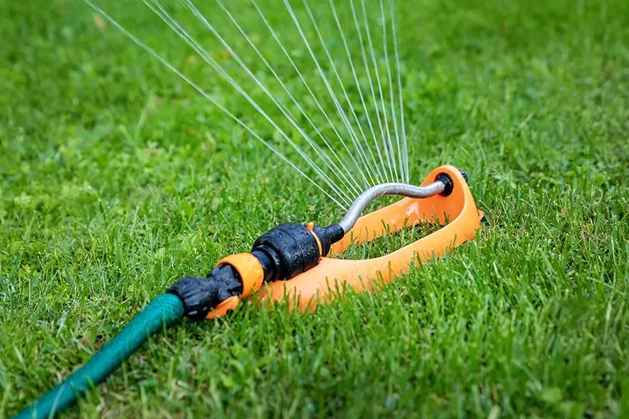 An bright orange oscillating lawn sprinkler attached to a green garden hose actively watering a yard.