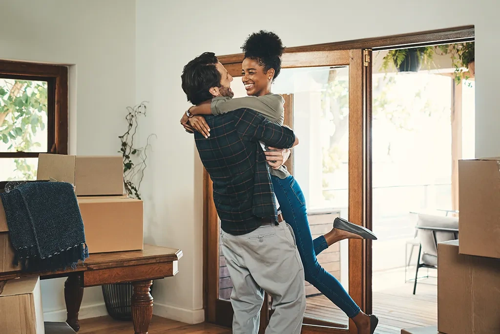 A happy man lifting and hugging a smiling woman inside a home surrounded by cardboard moving boxes.